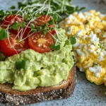 Avocado toast topped with cherry tomatoes and cottage cheese, served with scrambled eggs on a plate.