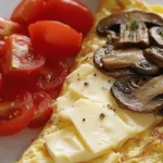 Mushroom omelette with melted Swiss cheese and fresh tomato slices on a white plate.