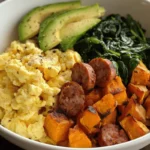 A hearty breakfast bowl featuring roasted sweet potatoes, seared sausage slices, scrambled eggs, sautéed greens, and fresh avocado, served in a white bowl.