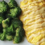 A fluffy cheesy omelette drizzled with creamy sauce, served with steamed broccoli on a white plate.
