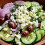 A Mediterranean cucumber salad with feta, olives, red onion, avocado, and fresh dill in a wooden bowl.