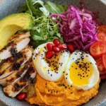 A healthy nourish bowl with poached eggs, spiced chicken, avocado, roasted veggies, and hummus.