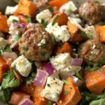 A vibrant salad with roasted sweet potatoes, meatballs, feta cheese, arugula, and red onions, garnished with fresh herbs.